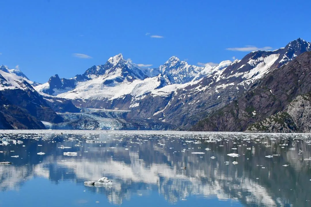 A glacier meeting still water beneath a snow-covered mountain range, with broken ice floating on the surface.